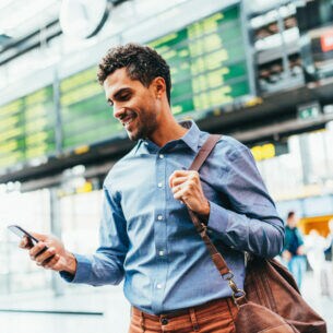 Ein junger Mann in blauem Hemd steht vor einer Abflugtafel am Flughafen. Er trägt eine Umhängetasche über der linken Schulter und schaut lächelnd auf das Smartphone in seiner rechten Hand