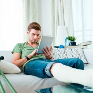 Eine Person mit gebrochenem Bein sitzt auf einem Sofa und schaut auf sein Tablet, neben ihr stehen Krücken.