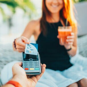 Eine Person in einer Hängematte hält ein Getränk in der Hand und bezahlt mit einer Kreditkarte.