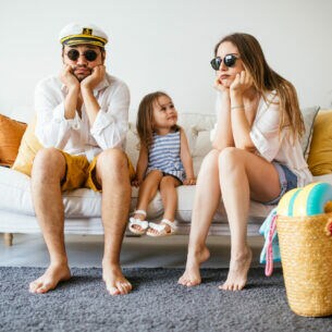 Eine Familie sitzt auf einem Sofa und stützt die Köpfe auf ihre Hände, daneben stehen gepackte Koffer.