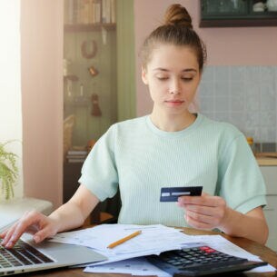 Eine Person sitzt am Küchentisch, schaut auf ihre Bankkarte und hat mehrere Dokumente vor sich liegen sowie die andere Hand auf einem geöffneten Laptop.
