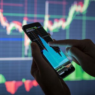 Nahaufnahme von zwei Händen, die ein Smartphone mit Aktienkurs halten. Im Hintergrund ist ein großes Display mit weiteren Kursen zu erkennen.