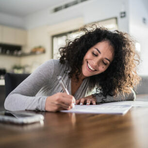 Eine Person sitzt am Tisch und trägt lächelnd etwas in ein Dokument ein.