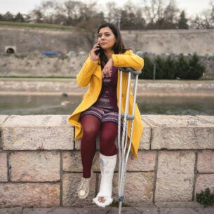 Eine Person mit eingegipstem Bein und Krücken sitzt auf einer Mauer am Fluss und telefoniert.