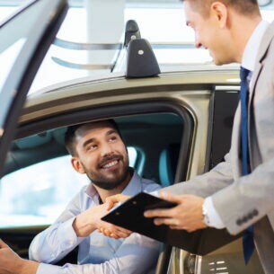 Eine Person sitzt im Autohaus in einem Auto und reicht einer anderen Person, die vor dem Auto steht, die Hand.