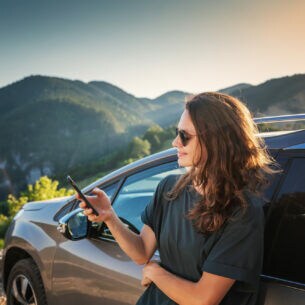 Eine Person lehnt an einem Auto und blickt auf ein Smartphone, im Hintergrund sind Berge zu erkennen.