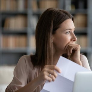 Eine Person sitzt vor einem Laptop, hält ein Blatt Papier in der Hand und blickt seitlich aus dem Bild.
