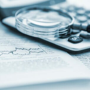 Nahaufnahme einer Lupe, die auf einem Taschenrechner und einem Papier mit Wirtschaftsinformationen liegt.