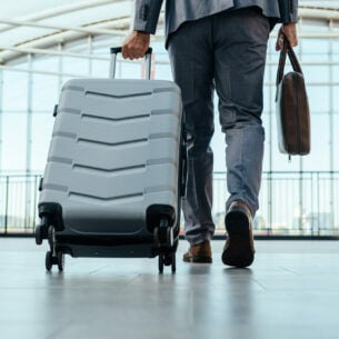 Ein Mann im Anzug geht mit einem Rollkoffer und einer Laptoptasche durch eine Halle.