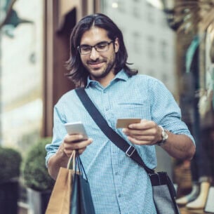 Eine Person mit Einkaufstüten steht vor einem Geschäft und hält Smartphone sowie eine Bankkarte in den Händen.