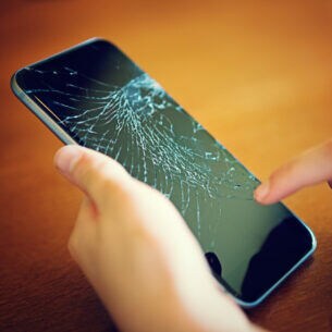 Eine Person hält ein Handy mit zersprungenem Display in der einen Hand und tippt mit einem Finger der anderen Hand darauf.