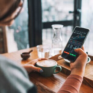 Eine Person sitzt im Café an einem Tisch und blickt auf ihr Smartphone, auf dem Aktienkurse zu sehen sind.