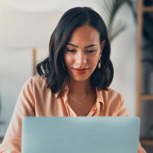 Eine lächelnde Frau sitzt an einem Tisch vor einem aufgeklappten Laptop.