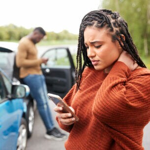 Zwei Autos stehen hintereinander auf einer Straße. Ein Mann lehnt am hinteren Auto, im Vordergrund sieht eine junge Frau besorgt auf ihr Smartphone.