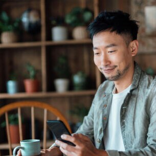 Eine Person sitzt an einem Tisch mit einer Kaffeetasse in der Hand und schaut auf ihr Smartphone in der anderen Hand.