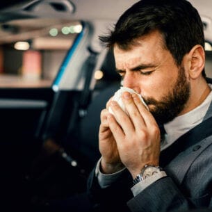 Ein dunkelhaariger Mann im Anzug sitzt angeschnallt am Steuer eines parkenden Autos und putzt seine Nase.