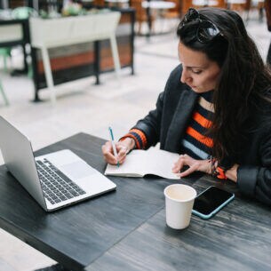 Eine Frau sitzt an einem Tisch vor einem aufgeklappten Laptop und schreibt mit einem Stift in ein geöffnetes Notizbuch.