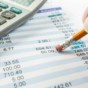 Eine Person hält einen Bleistift mit dem Radiergummi-Ende an eine Zahl auf einem Dokument. Auf dem Dokument liegt zudem ein Taschenrechner.