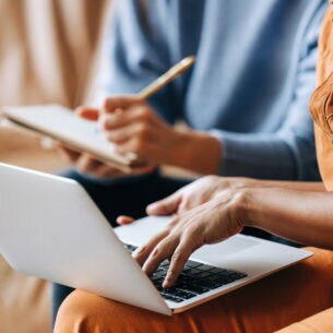 Eine Person hat einen Laptop auf dem Schoß und tippt auf der Tastatur, eine andere Person sitzt mit Notizblock und Stift daneben.