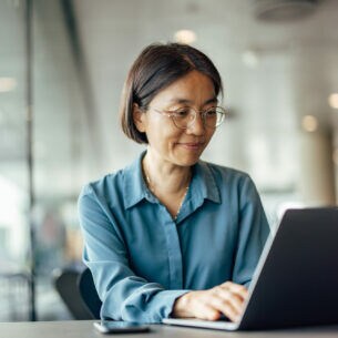 Eine Person sitzt lächelnd vor einem Laptop am Tisch.