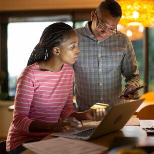 Eine Frau sitzt in einem Wohnzimmer mit einer Bankkarte vor einem aufgeklappten Laptop. Neben ihr steht ein Mann mit einem Handy in der Hand.