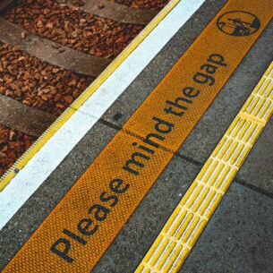 Der Sicherhinweis Please mind the gap auf dem Boden an einem Gleis.