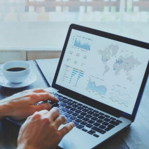 Eine Person hat die Hände auf der Tastatur eines ausgeklappten Laptops, neben dem eine Tasse Kaffee steht. Der Bildschirm zeigt eine Weltkarte und Kursverläufe.