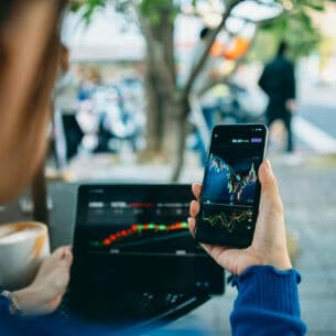 Eine Person im Freien an einem Tisch hält ein Smartphone in der Hand, auf dessen Display ein Börsenkurs gezeigt wird. Auf dem Tisch steht eine Kaffeetasse neben einem Notebook mit Kursdaten auf dem Bildschirm.