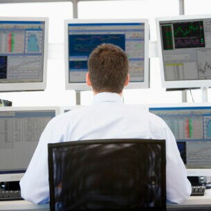 Eine Person sitzt am Schreibtisch vor sechs PC-Monitoren, auf denen Tabellen und Kursverläufe zu sehen sind.