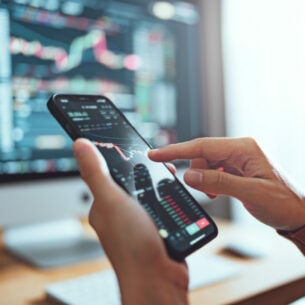 Eine Person hält ein Smartphone in der Hand. Auf dem Bildschirm ist ein Aktienkurs zu sehen. Im Hintergrund ein Computerbildschirm, der ebenfalls Kurse zeigt.