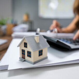 Eine Person sitzt an einem Tisch und tippt in einen Taschenrechner. Auf dem Tisch liegt ein Stapel Papier und steht ein Miniaturhaus.