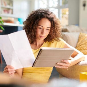 Eine Frau sitzt auf dem Boden neben einem Sofa. Sie hält in einer Hand ein Tablet und in der anderen ein Blatt Papier.