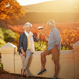 Ein älterer Mann und eine ältere Frau stoßen an eine Mauer gelehnt vor herbstlichen Laubbäumen mit einem Glas Rotwein an.