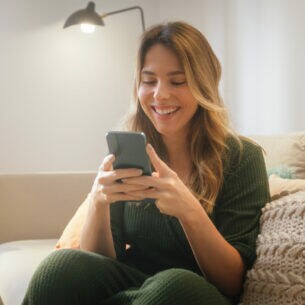 Eine Person sitzt auf einem Sofa und schaut lachend auf das Smartphone, das sie mit beiden Händen festhält.