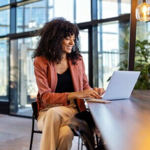 Eine Person sitzt in einem lichtdurchfluteten Ambiente an einem hochbeinigen Tisch und arbeitet am Laptop.