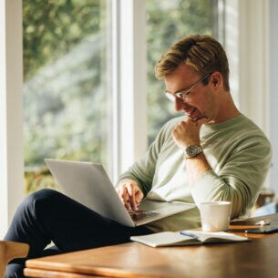 Eine Person sitzt am Esstisch und hat einen geöffneten Laptop auf dem Schoß.