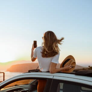 Eine Person in der Rückansicht schaut bei Sonnenuntergang aus dem Dach eines Autos und fotografiert sich dabei.