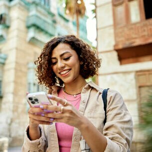 Eine Person steht zwischen hohen Steingebäuden und blickt auf das Smartphone in ihren Händen.
