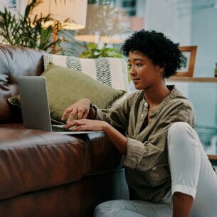 Eine junge Frau sitzt auf dem Boden neben dem Sofa und benutzt ein Notebook.
