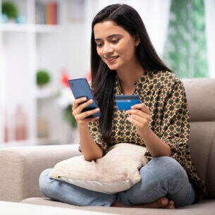 Eine Person sitzt im Schneidersitzt auf einem Sofa und hält sowohl eine Kreditkarte als auch ein Smartphone in den Händen.