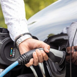Eine Person steht neben einem Auto mit geöffneter Tankklappe und hält das Ladekabel einer Wallbox in der Hand