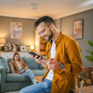 Eine Person sitzt auf einem Tisch und hält eine Kreditkarte sowie ein Smartphone in den Händen. Im Hintergrund sitzt eine weitere Person auf einem Sofa.