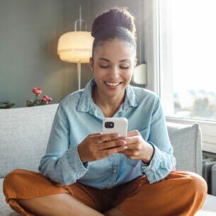 Eine Person sitzt im Schneidersitz auf einem Sofa und blickt auf ihr Smartphone, das sie in beiden Händen hält.