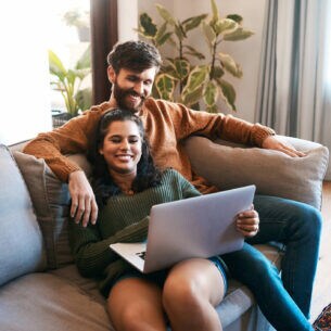 Zwei Personen sitzen gemeinsam auf einem Sofa und blicken in ein geöffnetes Notebook.