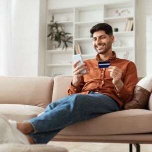 Eine Person sitzt mit ausgestreckten Beinen auf einem Sofa und hält eine Kreditkarte sowie ein Smartphone in den Händen.