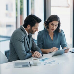 Zwei Personen in Businesskleidung sitzen an einem weißen Tisch, vor sich Kaffeetassen, ein Tablet und diverse Dokumente.