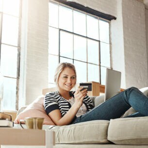 Eine Person liegt auf einem Sofa, hat ein Laptop auf ihrem Schoß und blickt lächelnd auf eine Bankkarte.