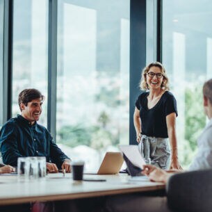 Vier Personen sitzen an einem Bürotisch, vor zwei Personen befindet sich ein aufgeklappter Laptop. An einer Tischseite steht eine lächelnde Frau, die den Blick zur Gruppe gewendet hat.