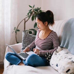 Eine Person sitzt im Schneidersitz auf einem Sofa mit aufgeklapptem Notebook auf dem Schoß.