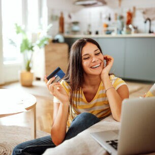 Eine Person sitzt auf dem Fußboden zwischen Couchtisch und Sofa, hält eine Kreditkarte in der Hand und blickt auf ein geöffnetes Notebook, das auf der Sofasitzfläche steht. 